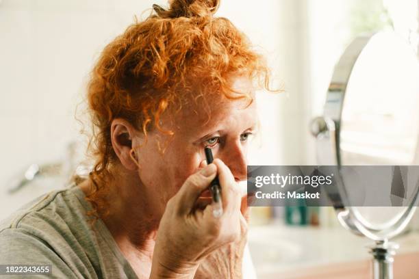 senior woman looking in mirror and applying eyeliner at home - kajalschminke stock-fotos und bilder