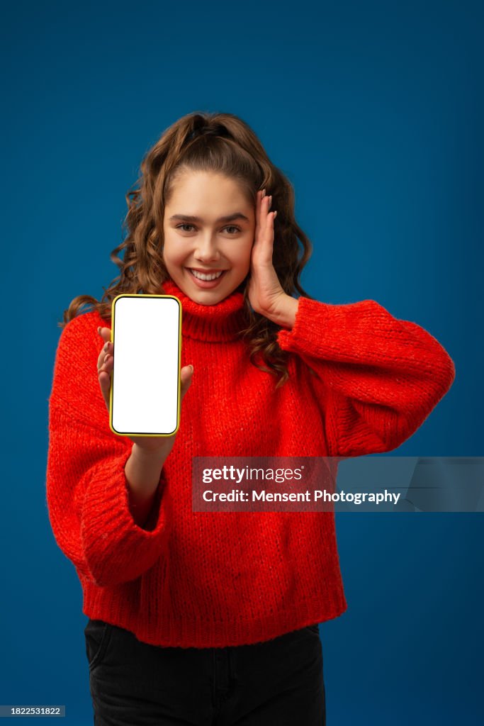 Amazed beautiful girl holding smartphone mockup with a white screen in blue background
