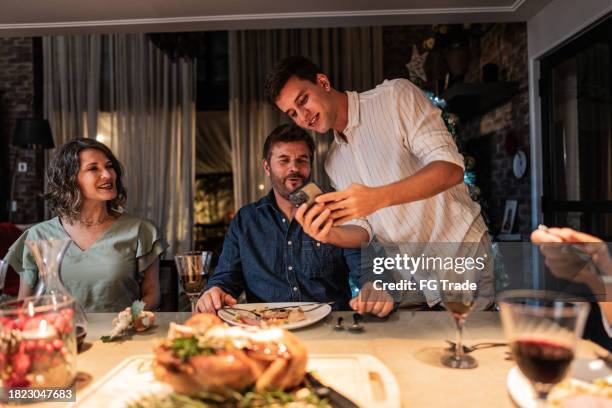 son showing something on the mobile phone to his parents at christmas dinner - zoom dinner stock pictures, royalty-free photos & images