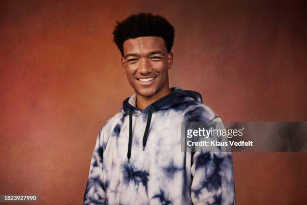 portrait of smiling teenage boy in tie dye hooded shirt standing against brown background - one teenage boy only stock pictures, royalty-free photos & images