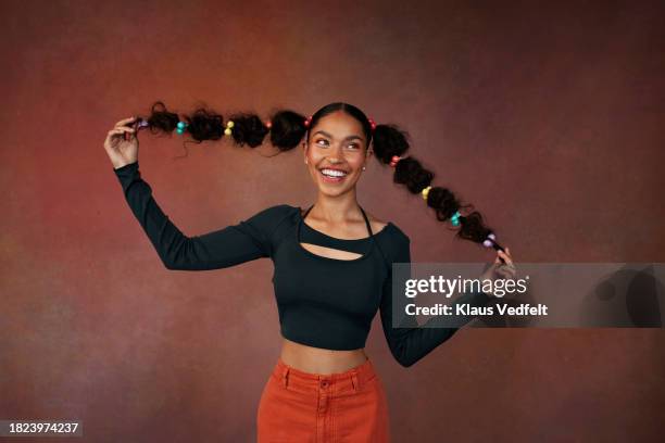 happy teenage girl holding braided hair while standing against brown background - braid hairstyle stock pictures, royalty-free photos & images
