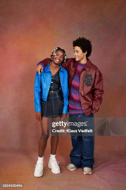 teenage boy arm around with female friend standing against brown backdrop - hands in pockets stock pictures, royalty-free photos & images