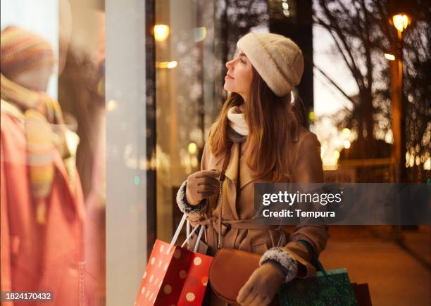 christmas window scrutiny: a waist-up view captures the woman in her thirties, dressed in winter clothes, as she attentively examines the christmas shop display adorned with lights - boutique stock pictures, royalty-free photos & images