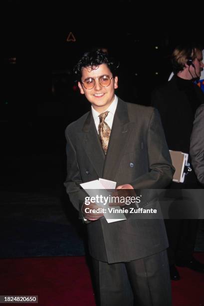 American actor Max Casella wearing a double-breasted suit with a white shirt and a brown-and-gold tie, and glasses with circular lenses, holding a...