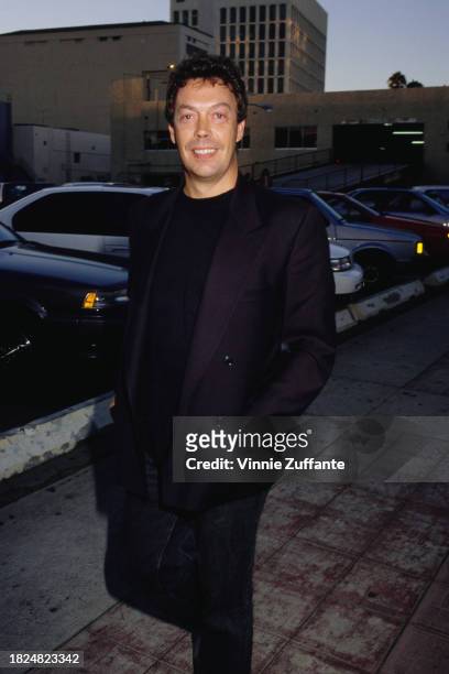 British actor and singer Tim Curry wearing a black jacket over a black crew neck top, walking beside a parking lot, United States, circa 1992.