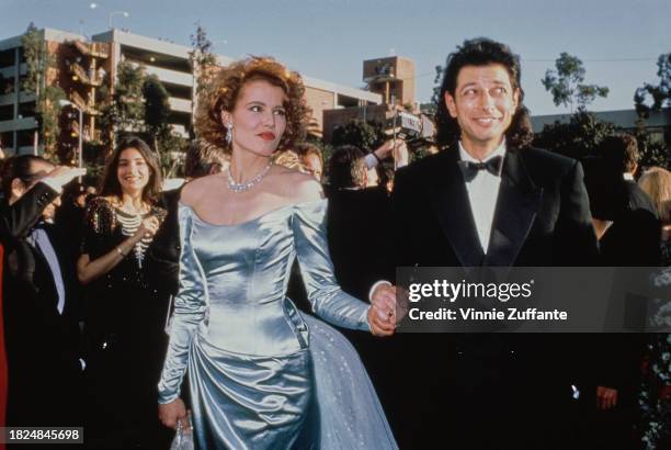 American actress Geena Davis wearing an off-shoulder blue evening gown, holding hands with her husband, American actor Jeff Goldblum, who wears a...