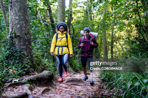 couple of hikers walking through the forest. young man with a disability and using a prosthesis on his leg trekking with his girlfriend or friend. concept of dignity, inclusion, empowerment, trust, community, authenticity, diversity and disability. - senderismo fotografías e imágenes de stock