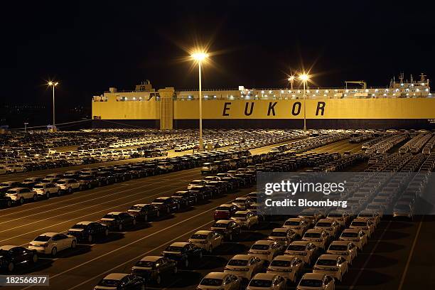 Kia Motors Corp. Vehicles bound for export await shipment in front of a Eukor Car Carriers Inc. Roll-on/roll-off cargo ship at night at the port of...