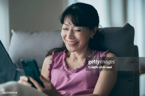 im wohnzimmer liegt eine frau mittleren alters gemütlich auf dem sofa, vertieft in ihr smartphone. mit gemächlichem wischen und tippen navigiert sie durch das internet und frönt möglicherweise dem online-shopping. - online dating stock-fotos und bilder