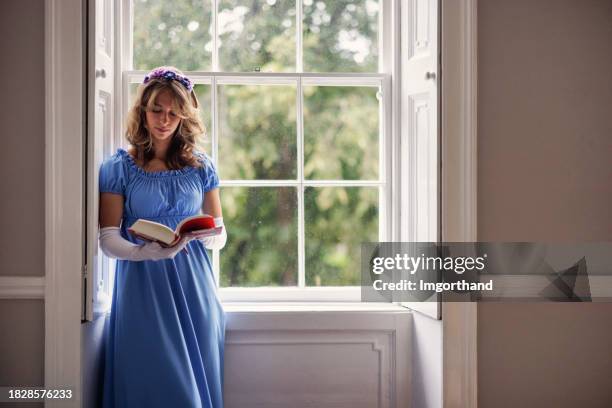 jovem mulher usando um vestido da era regência está lendo um livro perto de uma janela - estilo regency - fotografias e filmes do acervo