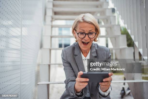 a mature woman is playing video games on her mobile phone while outside on a break from work - handheld video game stock pictures, royalty-free photos & images