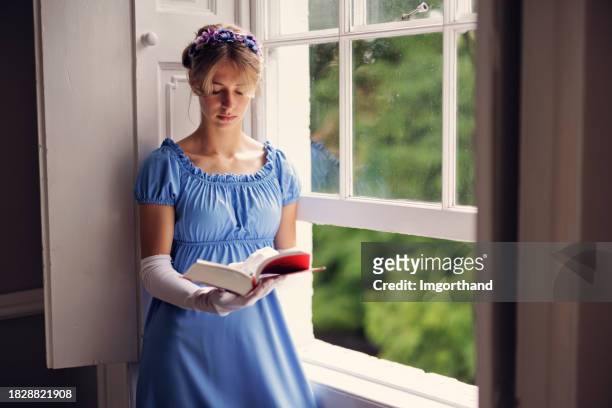 junge frau in einem kleid aus der regency-ära liest ein buch in der nähe eines fensters - girl dress romantic stock-fotos und bilder