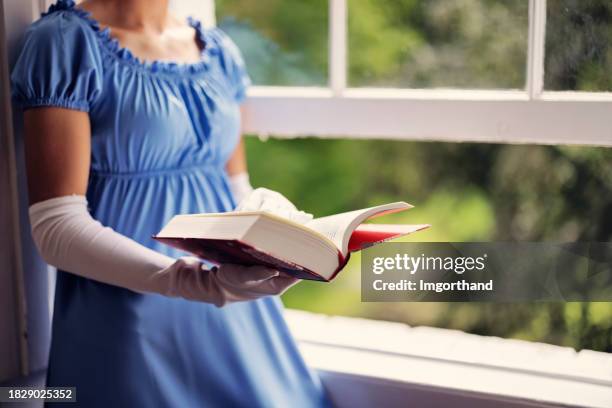 young woman wearing a regency era dress is reading a book near a window - material stock pictures, royalty-free photos & images