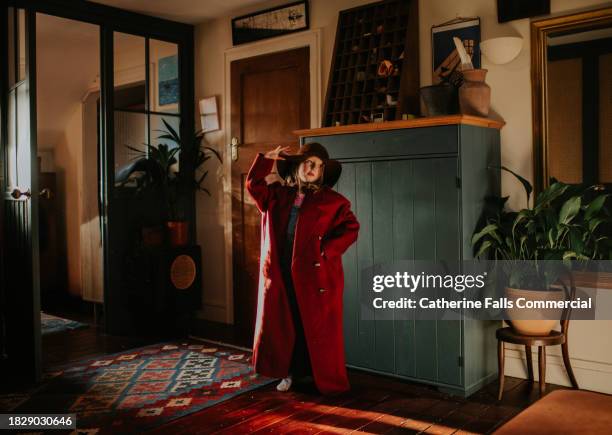 a young girl stands in a stylish bedroom, wearing an adult's coat that is too big and a wide brimmed hat - childrens closet stock-fotos und bilder