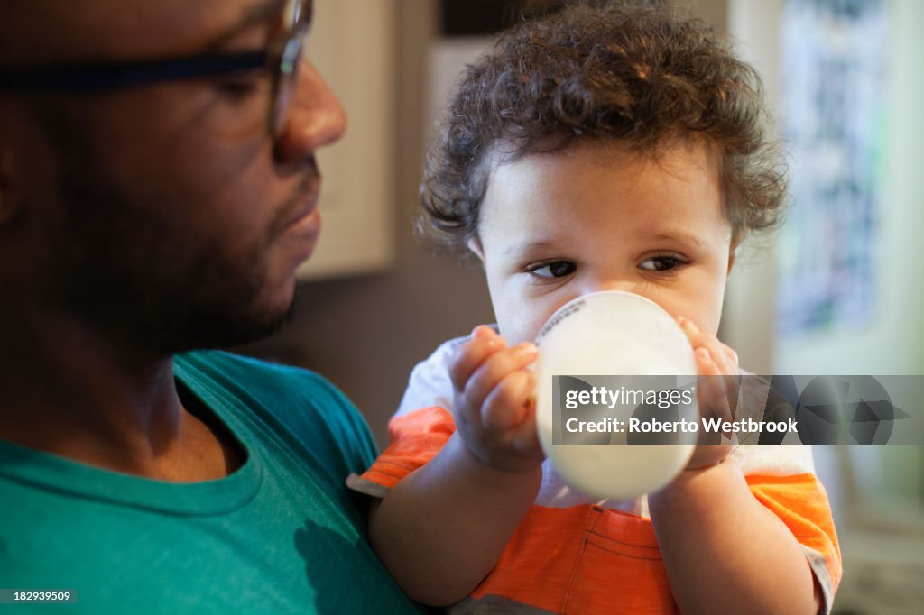 Father helping baby drink bottle in kitchen