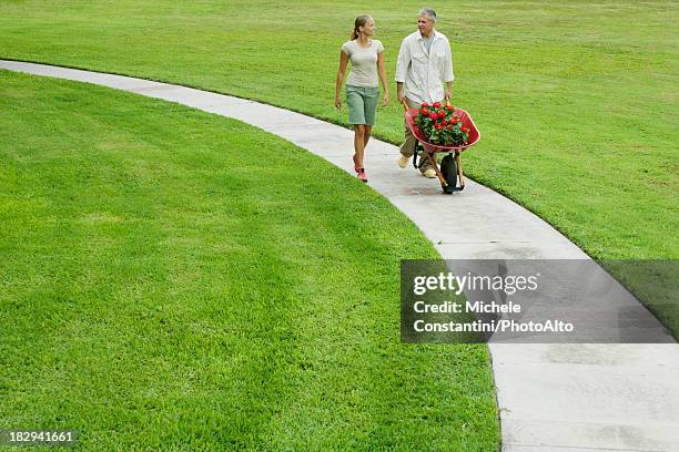 couple walking along footpath, man pushing wheelbarrow filled with flowers - sentiero di giardino foto e immagini stock