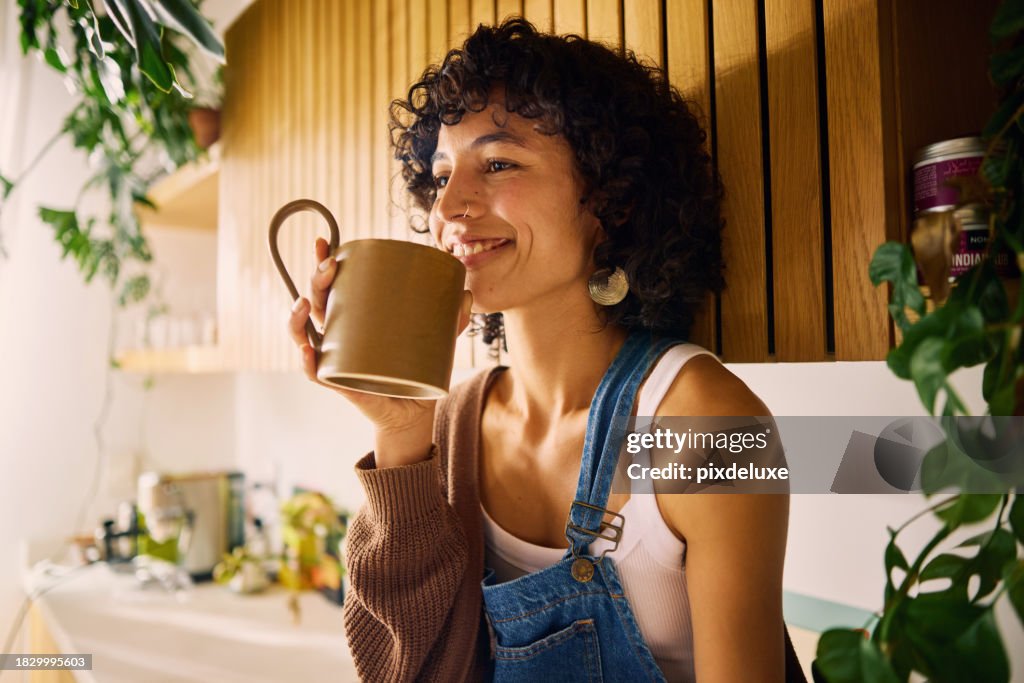 Zufriedene Frau, die das Aroma ihres Morgenkaffees in einer gut beleuchteten Küche mit modernen Holzakzenten und üppigen Zimmerpflanzen genießt, einen Moment der Gelassenheit umarmt und Achtsamkeit und die Freude an einfachen Freuden im Alltag symbolisi