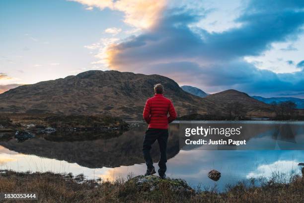 rear view of a man in a red puffer coat looking at the sunset view of a loch in the scottish highlands - laje stock pictures, royalty-free photos & images