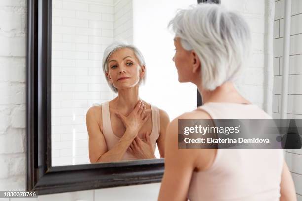elderly happy woman admires herself looking in mirror while standing in bathroom against white background - hals stock-fotos und bilder