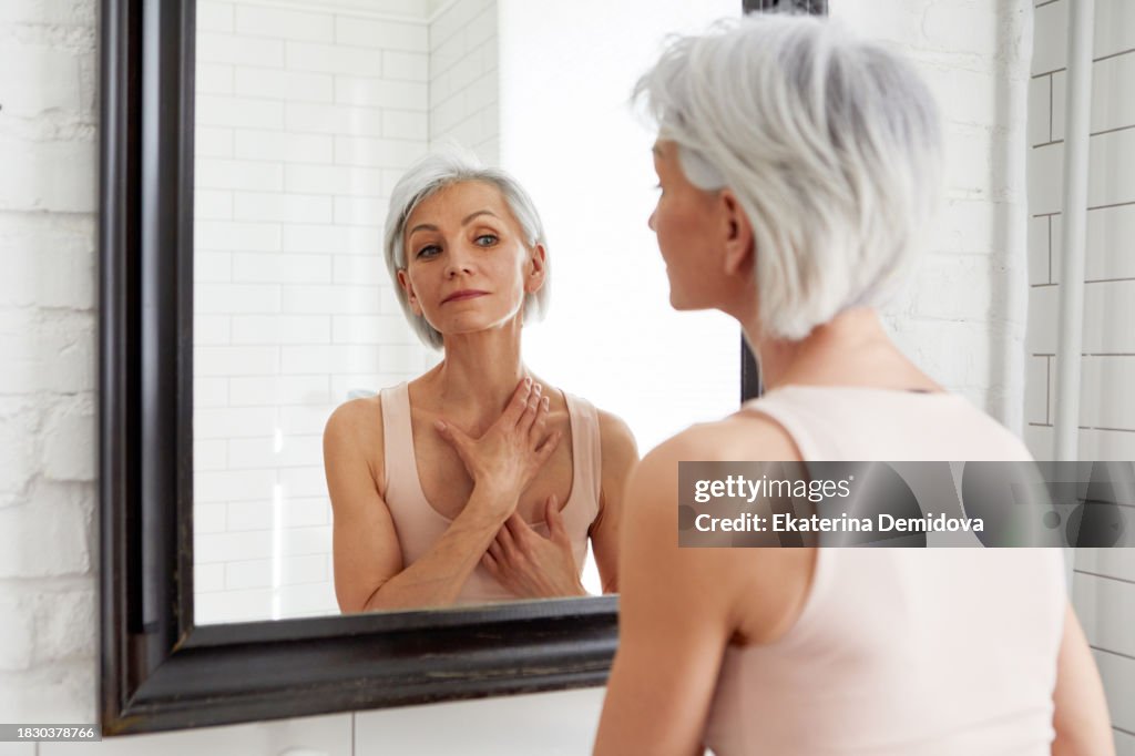 Elderly happy woman admires herself looking in mirror while standing in bathroom against white background