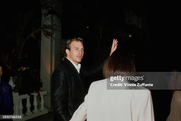 American actor and film director Kevin Costner, wearing a black jacket over a white shirt, turns to wave while with his wife, Cindy Silva, who has...