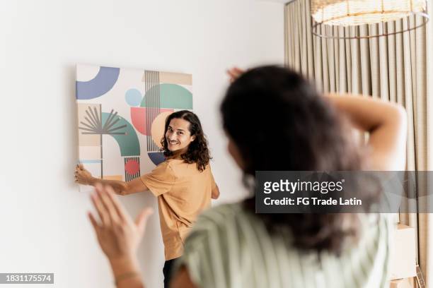 mid adult man asking his wife for help to put the frame on the wall in living room at new house - house for an art lover stock pictures, royalty-free photos & images