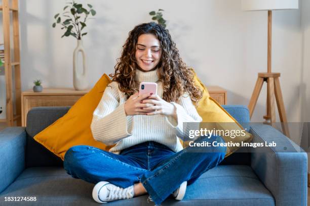 smiling young woman sitting on sofa and using mobile phone at home - mensagem com foto imagens e fotografias de stock