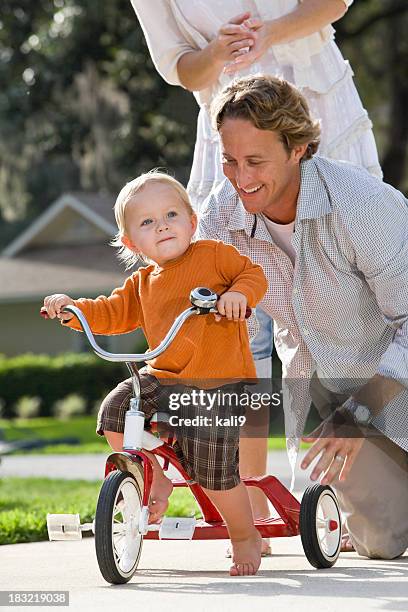 padre ayudando a toddler ride triciclo, madre standing behind - triciclo fotografías e imágenes de stock