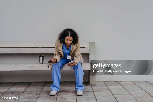 female doctor feeling overwhelmed at work, sitting on bench, smoking cigarette and drinking coffee. healthcare worker taking break from work, resting outdoors, feeling exhausted. - zigarettenpause stock-fotos und bilder