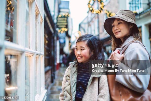 cheerful teenage girl looking at the window display of a gift shop with her mom while doing christmas shopping in high street of a town - retail equipment stock pictures, royalty-free photos & images