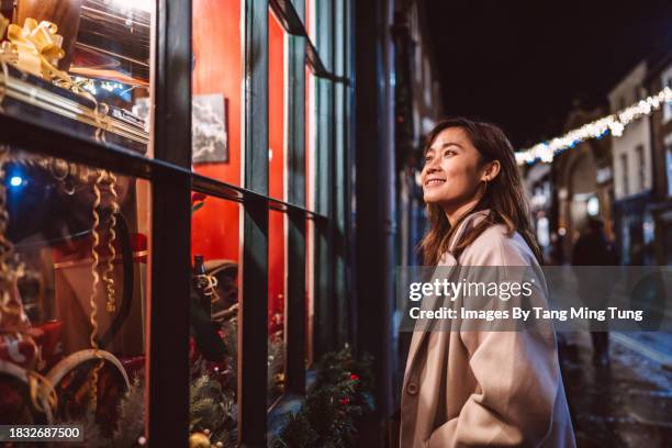 beautiful asian woman looking at the store window while doing christmas shopping in high street at night - commercial event stock pictures, royalty-free photos & images