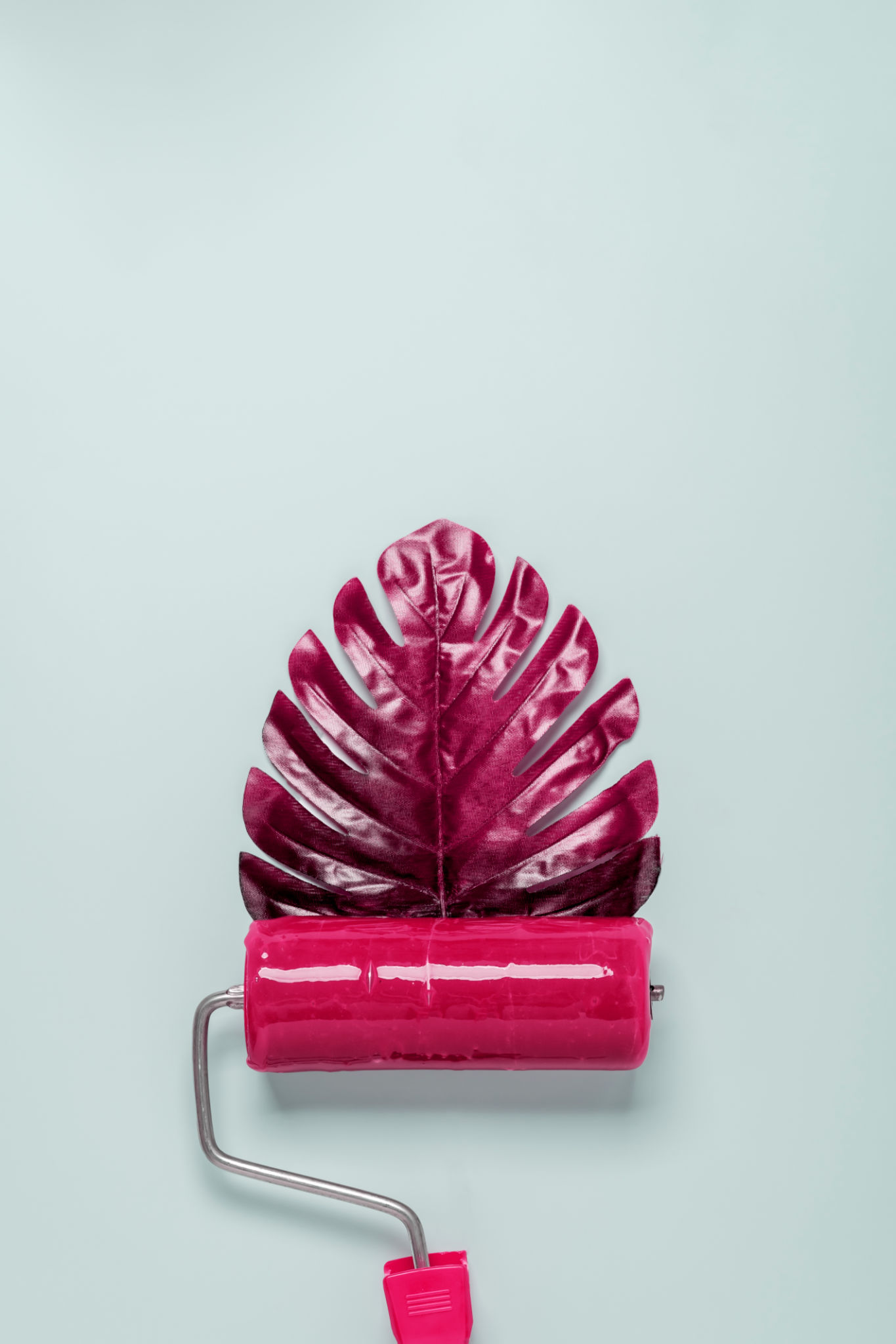 A paint roller with red paint creates a red monstera leaf on a pastel blue background. A paint roller with red paint creates a red monstera leaf on a pastel blue background.