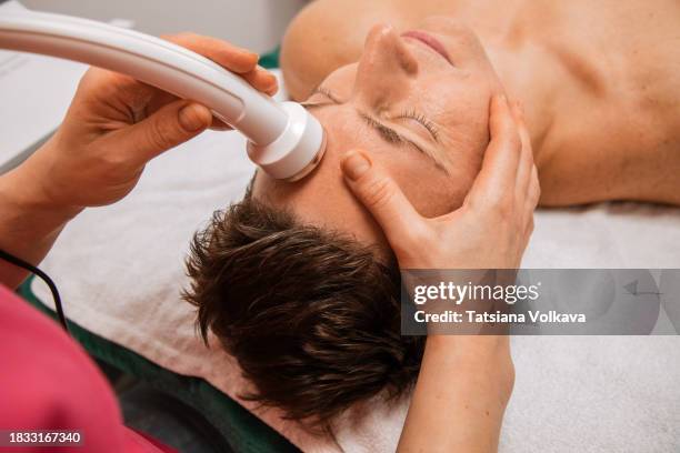 proficient beauty therapist in red uniform expertly administers ultrasound facial therapy with high-tech white device - administers stock pictures, royalty-free photos & images