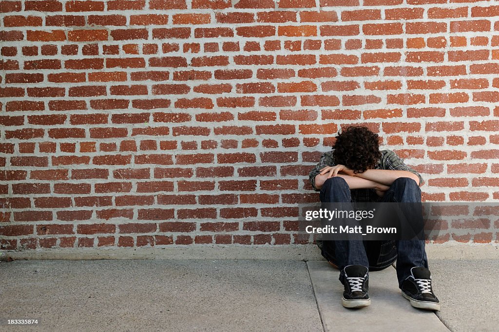 Lonely teenager sits against brick wall with arms cover eyes