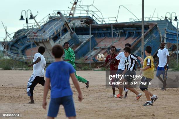 Italian and Eritrean migrant boys play football at Lampedusa Football Field where boats fomerly used by immigrants arriving in Lampedusa lie disused...