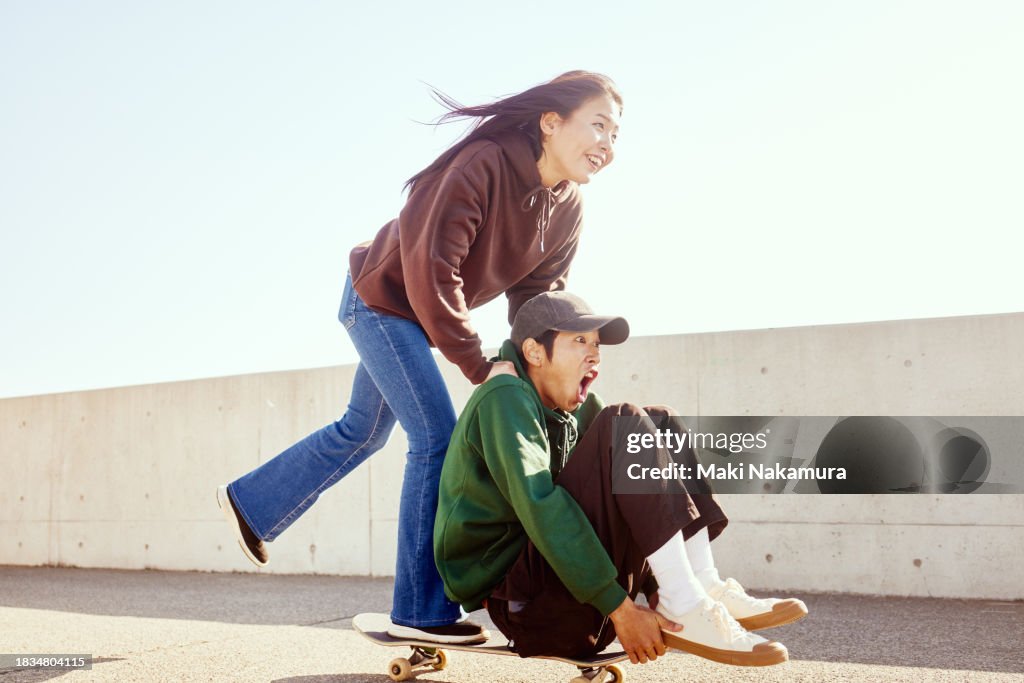 Young man enjoys pushing his friend sitting on a skateboard from behind with his hand.