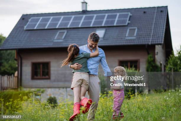 father and daughters having fun in front their family house with solar panels on the roof - zonnepanelen stockfoto's en -beelden