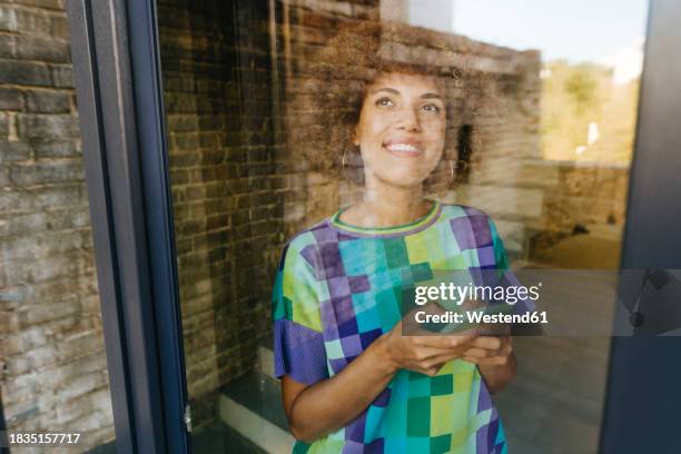 thoughtful woman holding smart phone seen through glass window - photographed through window stockfoto's en -beelden