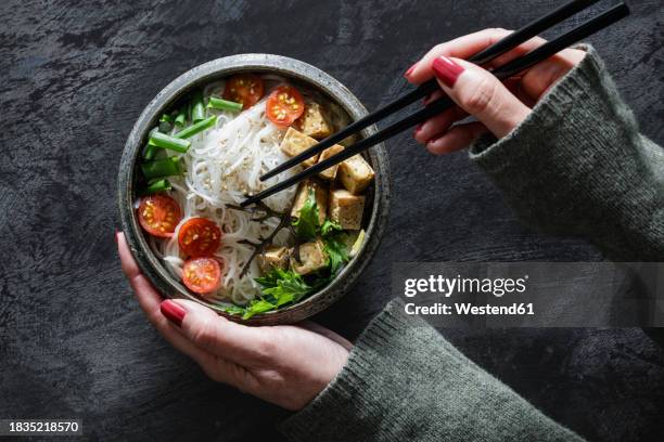 hands of woman eating bowl of vegan tomkhakaisoup with tofu, tomatoes, salad, rice noodles, sesame seeds and scallion - asiatische küche stock-fotos und bilder