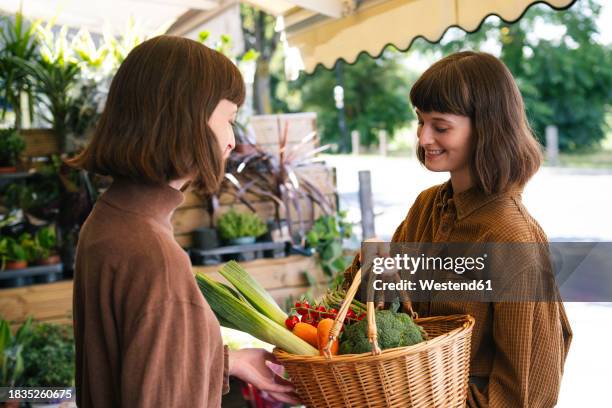 smiling twin sisters holding basket of vegetables at farmer's market - leek stock pictures, royalty-free photos & images