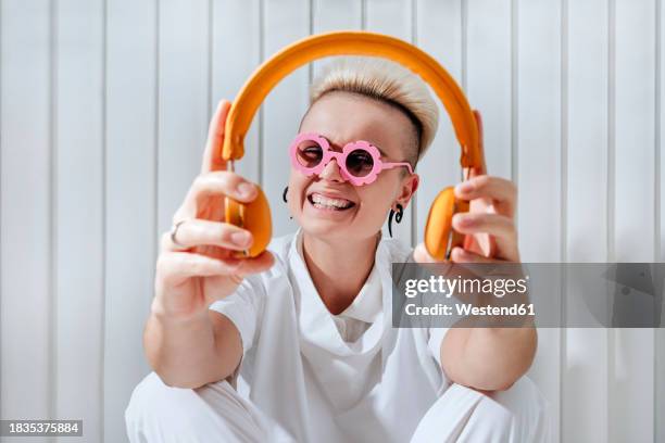 happy young woman wearing funky glasses and holding wireless headphones in front of wall - pompadour stock pictures, royalty-free photos & images