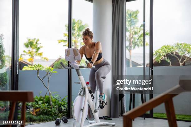 active woman exercising on exercise bike and using tablet pc at home - exercise bike stock pictures, royalty-free photos & images