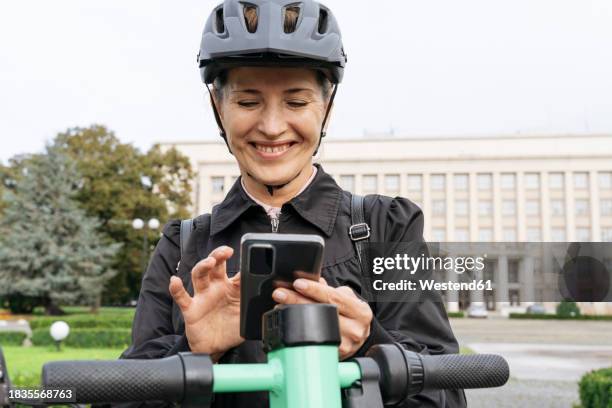 smiling woman standing with electric push scooter and using smart phone in park - cycling helmet stock pictures, royalty-free photos & images