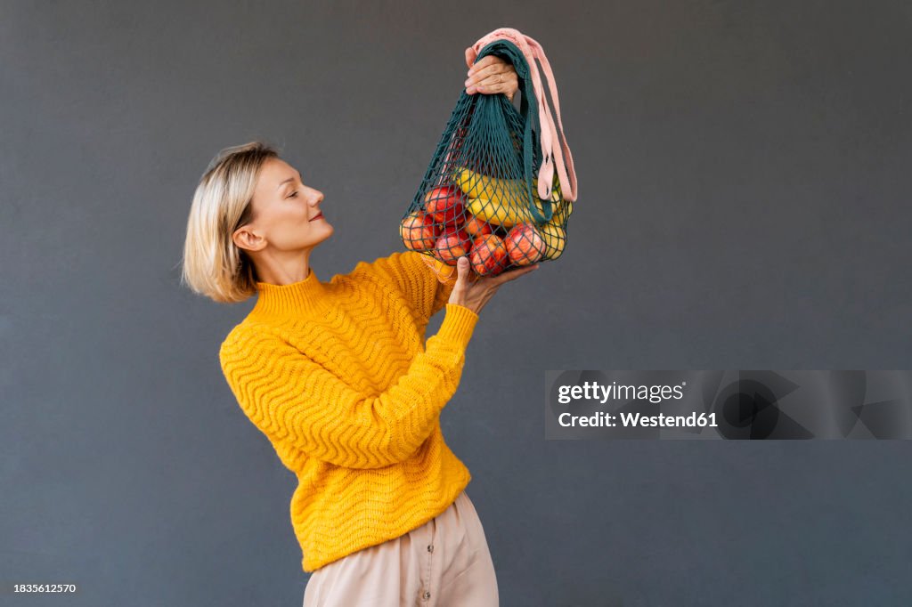 Smiling woman looking at mesh bags with fruits against black background