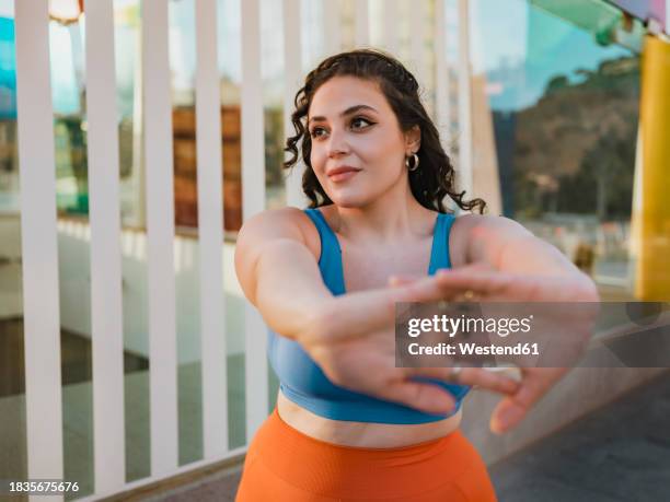young woman in sports clothing stretching arms near building - voluptuoso fotografías e imágenes de stock