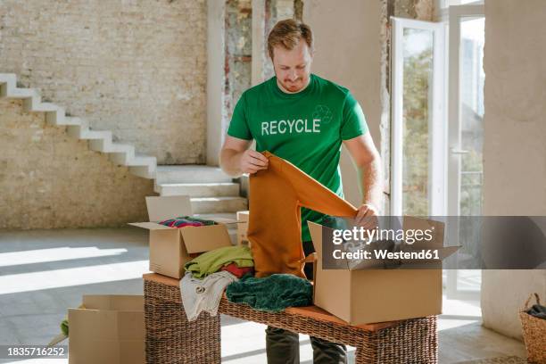 smiling activist folding secondhand clothes near donation box in apartment - man holding donation box stock pictures, royalty-free photos & images