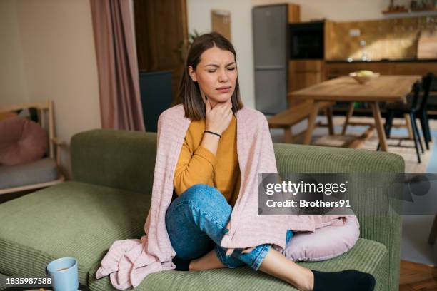 caucasian woman suffering from sore throat sitting on sofa - throat pain stock pictures, royalty-free photos & images