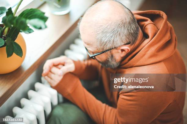 mature adult man warms himself by the radiator in the room - climate grief stock pictures, royalty-free photos & images