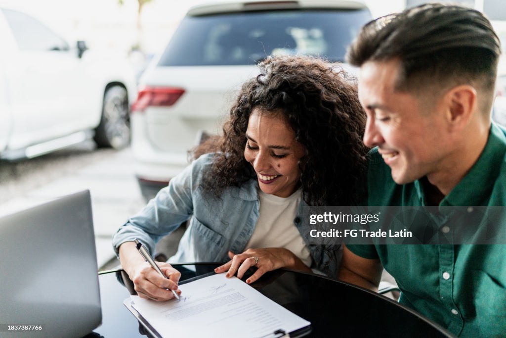 Mid adult couple signing documents to buy a car at dealership
