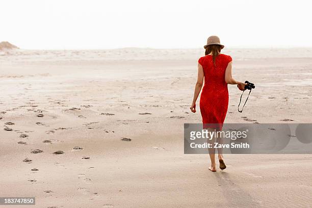 mature woman wearing red dress walking on beach - red dress stock-fotos und bilder